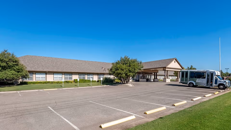 Exterior view of Brookdale Monterey senior living facility showing a single-story building with a covered entrance, a parking lot with marked spaces, a shuttle bus parked near the entrance, green grass, and trees under a clear blue sky.