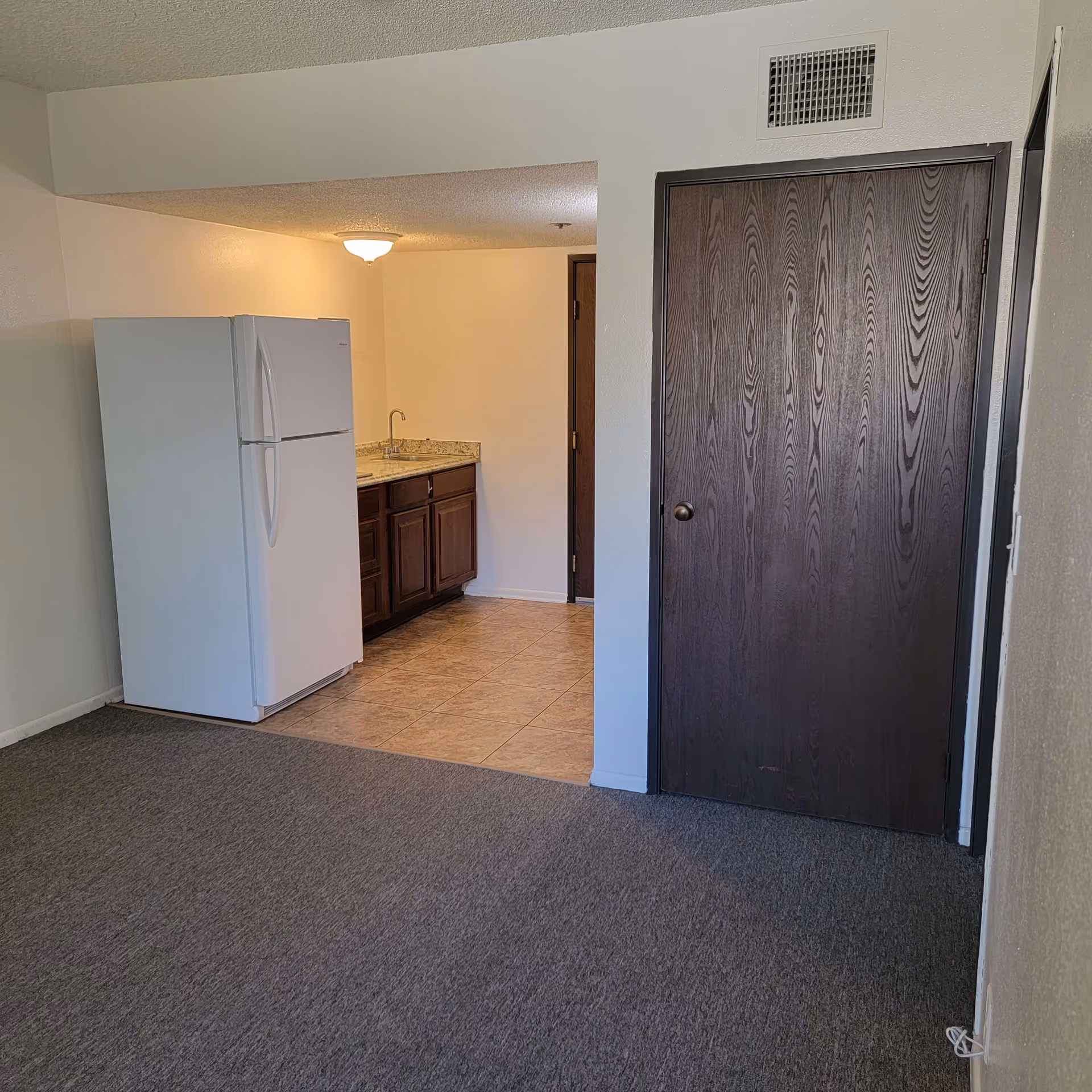 Interior view of a small kitchen area with a white refrigerator, dark wood cabinets, a granite countertop with a sink, and a tiled floor. Adjacent to the kitchen is a carpeted room with a dark wood door and white walls.