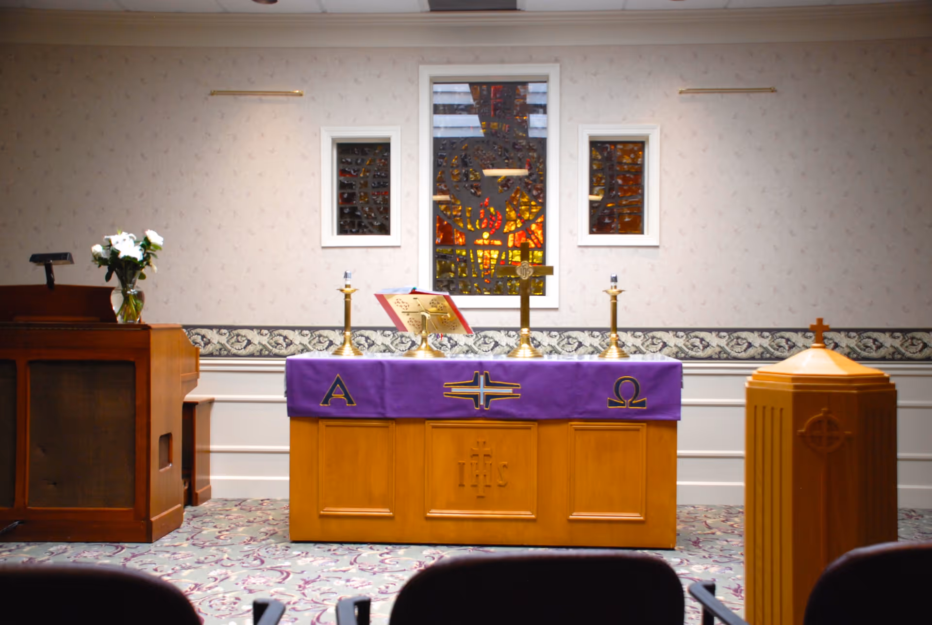 Interior view of a small chapel or worship room with a wooden altar covered by a purple cloth featuring Christian symbols Alpha and Omega. On the altar are two candlesticks, a cross, and an open book. Behind the altar is a stained glass window with religious imagery. To the left is a wooden lectern with a vase of white flowers, and to the right is a wooden baptismal font with a cross on top. The room has patterned carpet and wallpaper with a decorative border.