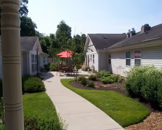 Outdoor walkway between single-story buildings with well-maintained landscaping, green grass, shrubs, and a patio area with a red umbrella and outdoor seating under a clear blue sky.