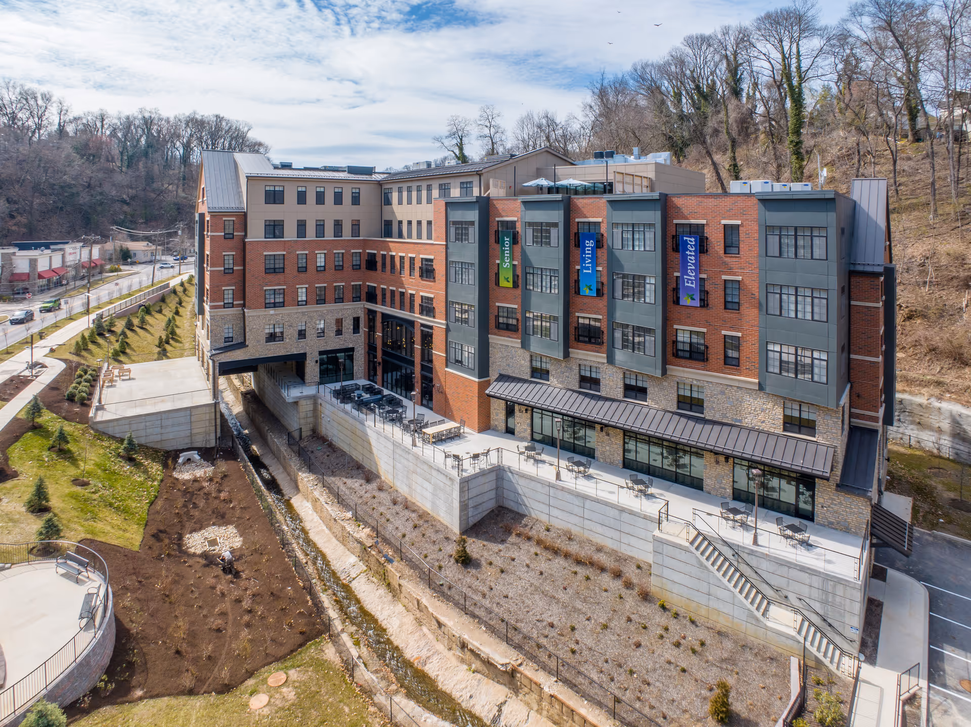 A multi-story senior living facility building with brick and stone exterior walls, large windows, and outdoor patio seating areas. The building is situated next to a small creek with landscaped grounds and trees in the background under a partly cloudy sky.