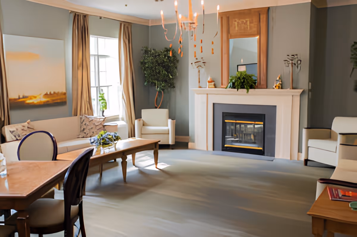 A cozy living room with a fireplace, a large mirror above the mantel, two armchairs, a sofa with cushions, a wooden coffee table, a dining table with chairs, and a large window with beige curtains letting in natural light. There are decorative plants and artwork on the walls.