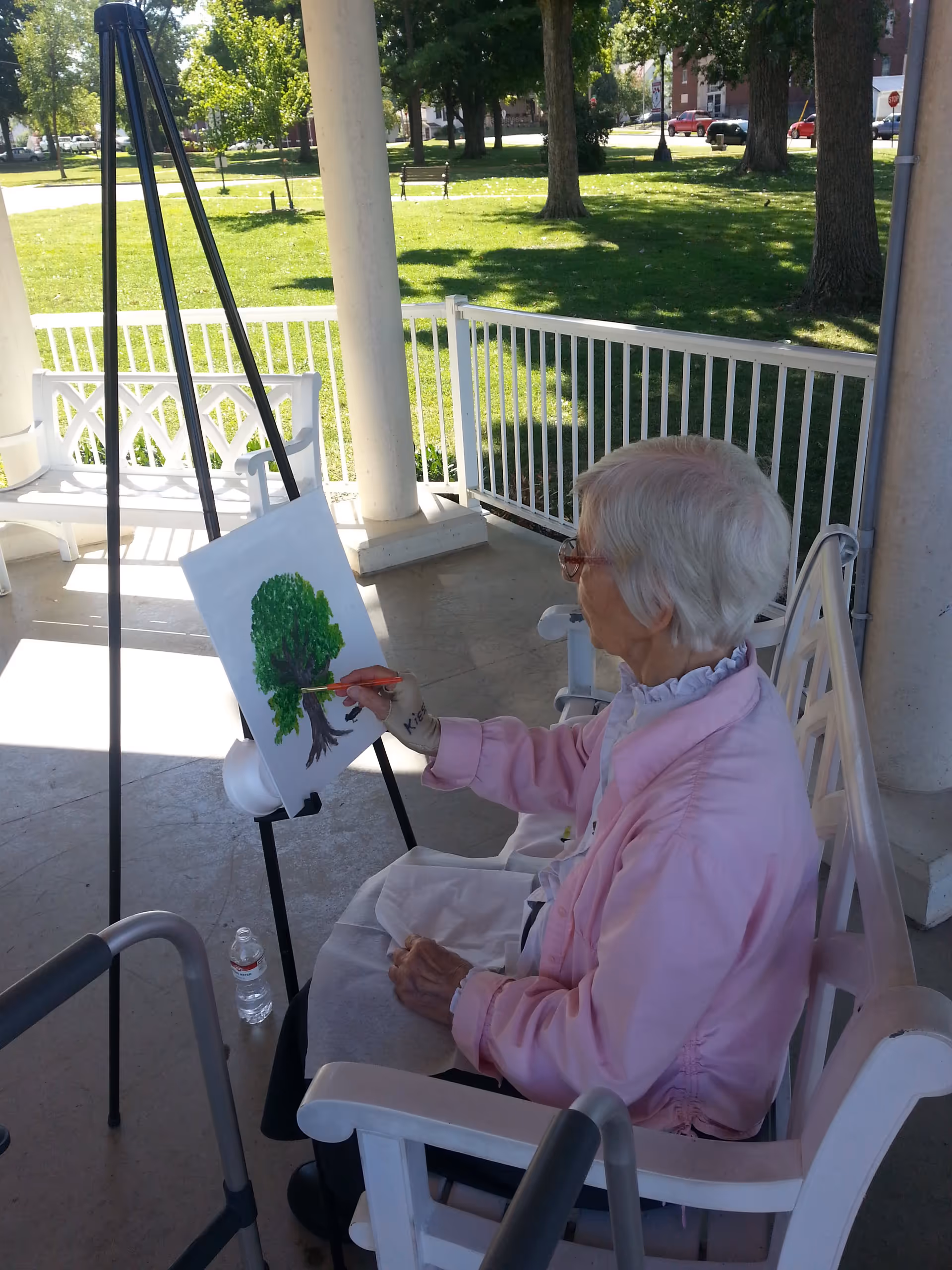 An elderly woman with white hair and glasses is sitting on a white bench on a covered porch, painting a green tree on a canvas set on an easel. She is wearing a pink jacket and has a walker nearby. The background shows a grassy area with trees and benches in a sunny outdoor setting.