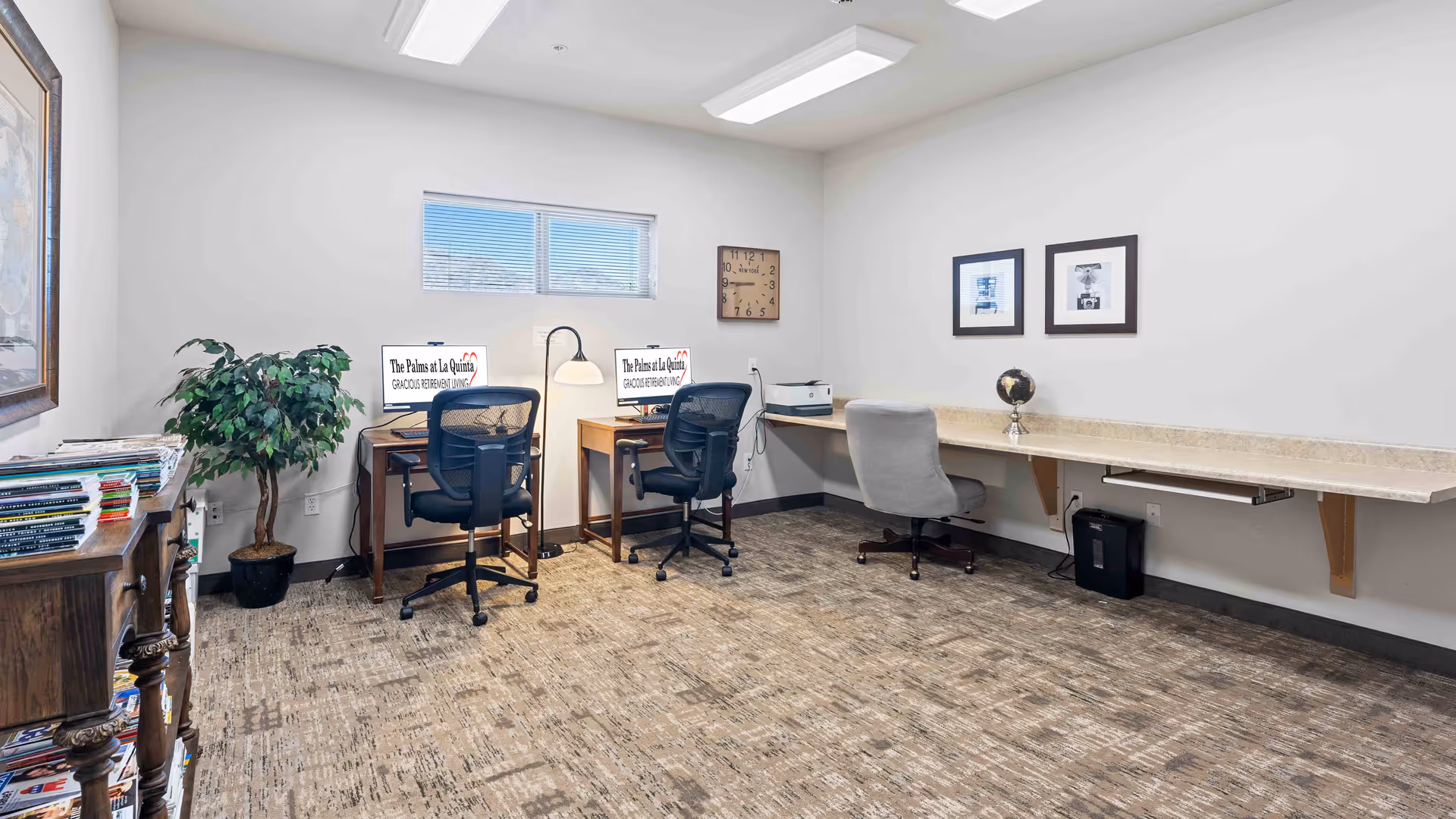 A well-lit office or computer room with two desks each equipped with a computer and office chair. There is a long wall-mounted counter with a gray office chair, a globe, and a printer. The room has a window, a potted plant, framed pictures on the wall, a clock, and a wooden bookshelf filled with magazines.