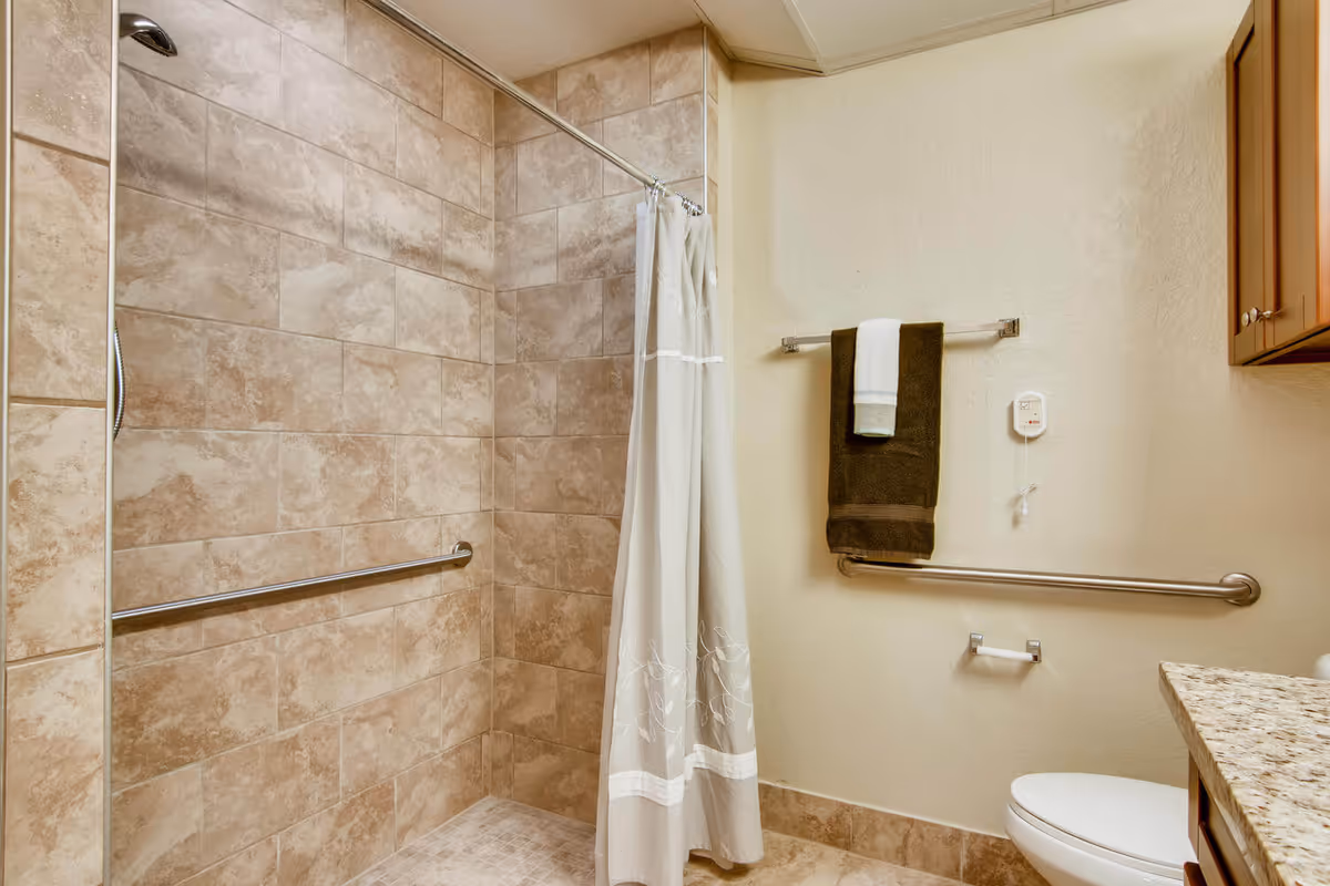A bathroom with beige tiled walls and floor, featuring a walk-in shower with a silver grab bar and a white shower curtain with a subtle leaf pattern. On the adjacent wall, there is a towel rack holding a dark brown towel with a white towel on top, a call button, and another silver grab bar below. A toilet and part of a granite countertop with wooden cabinets are visible on the right side.