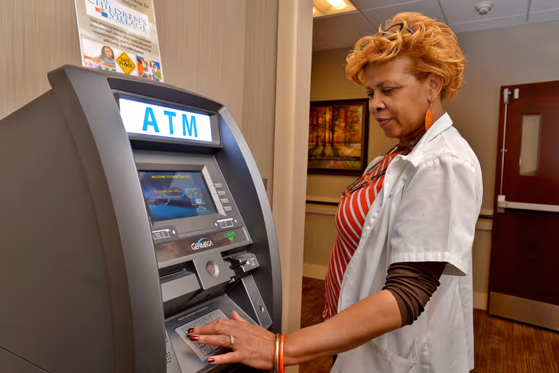 A woman with short curly hair wearing a white coat and striped shirt is using an ATM machine indoors. The ATM screen displays the word ATM in large blue letters. There is a poster on the wall above the ATM and a framed picture of a forest scene on the wall in the background.