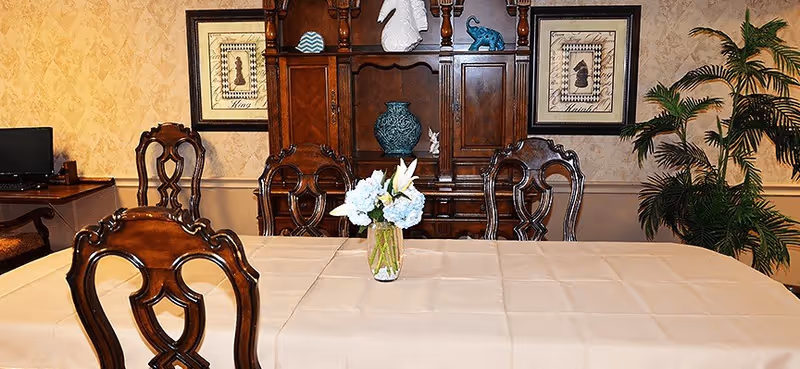 A dining room with a rectangular table covered with a beige tablecloth and a vase of white and blue flowers in the center. Surrounding the table are ornate wooden chairs. In the background, there is a wooden cabinet with decorative items and framed artwork on the wall. A green potted plant is visible on the right side.