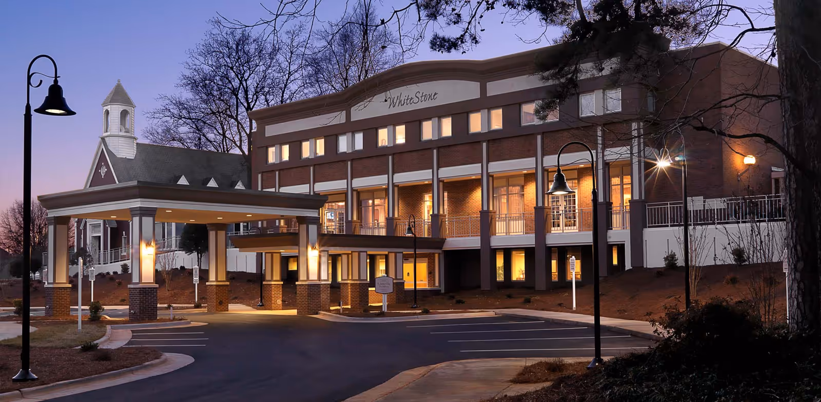 Front exterior of the WhiteStone senior living building at dusk showing a lit porte-cochère entrance, parking area, and street lamps.
