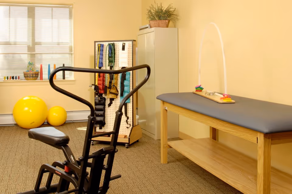 A therapy or exercise room with a black exercise machine in the foreground, two yellow exercise balls near a window with blinds, a pegboard with various colored straps and equipment, and a wooden therapy table with a gray cushion and a small colorful ring toss game on top.