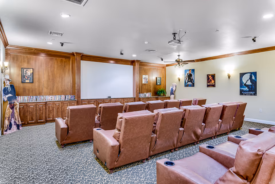 A senior living facility movie theater room with multiple rows of brown leather recliner chairs facing a large blank projection screen. The room has wood-paneled walls on one side with shelves holding DVDs, movie posters on the opposite wall, and a patterned carpeted floor. There are ceiling lights and a ceiling fan with a projector mounted above.