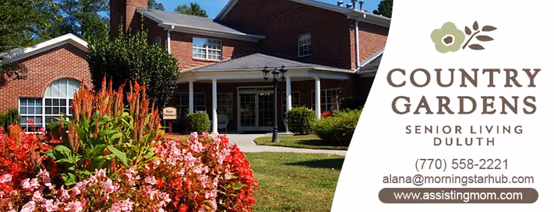 Exterior view of a red brick senior living facility building with a covered entrance, surrounded by a well-maintained garden featuring colorful flowers and greenery under a clear blue sky.