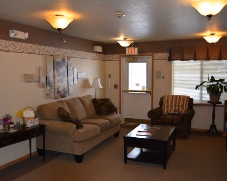 A cozy living room area with a beige sofa and patterned armchair. There is a dark wooden coffee table in front of the sofa with magazines on it. A side table with flowers and a basket is next to the sofa. The room has beige walls with a decorative border near the ceiling, ceiling lights, and a window with blinds and a valance. An exit door is visible in the background.