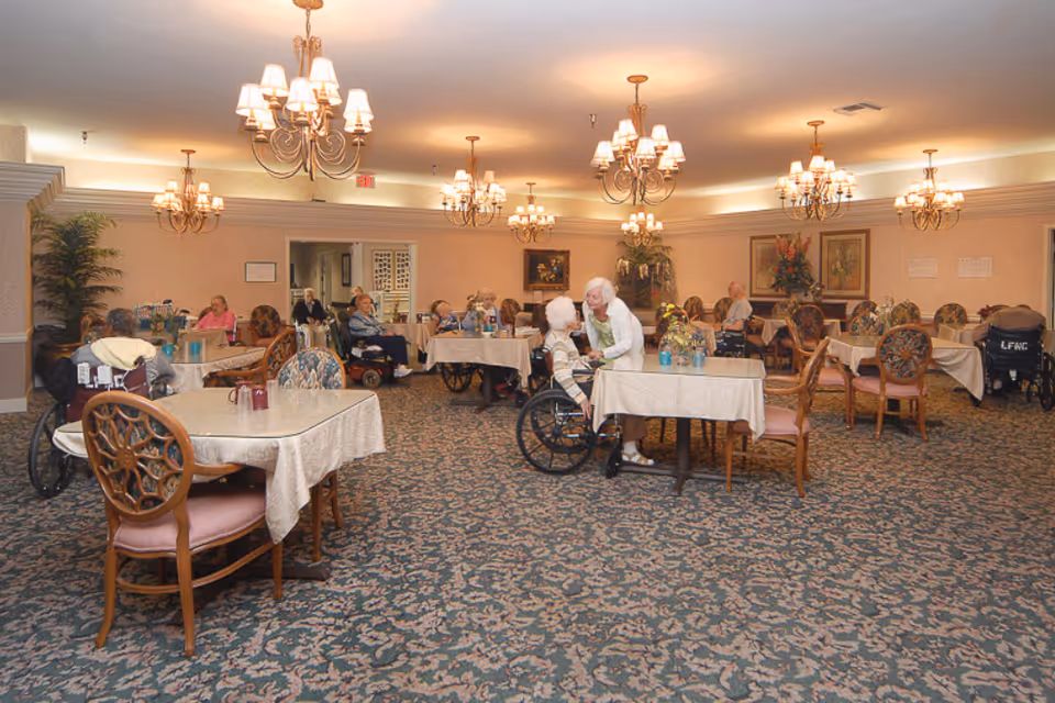 A spacious dining room in a senior living facility with several elderly residents seated at tables covered with white tablecloths. Some residents are in wheelchairs, and a caregiver is interacting with one of them. The room is decorated with chandeliers, framed artwork, floral arrangements, and has patterned carpet flooring.