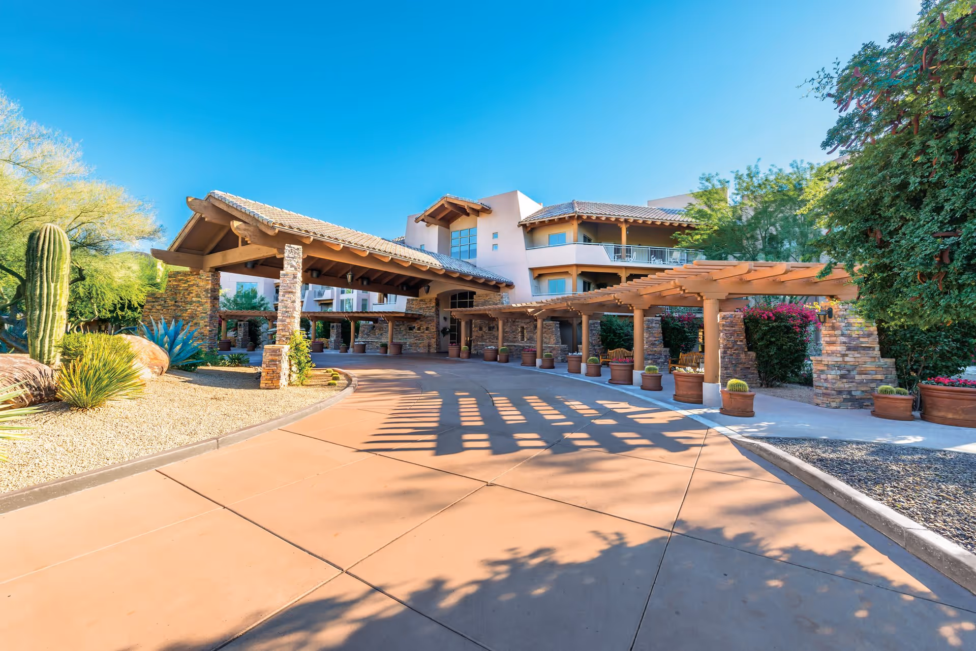 Exterior view of Vi at Grayhawk senior living facility entrance with a covered driveway, stone pillars, desert landscaping including cacti and shrubs, and a clear blue sky.