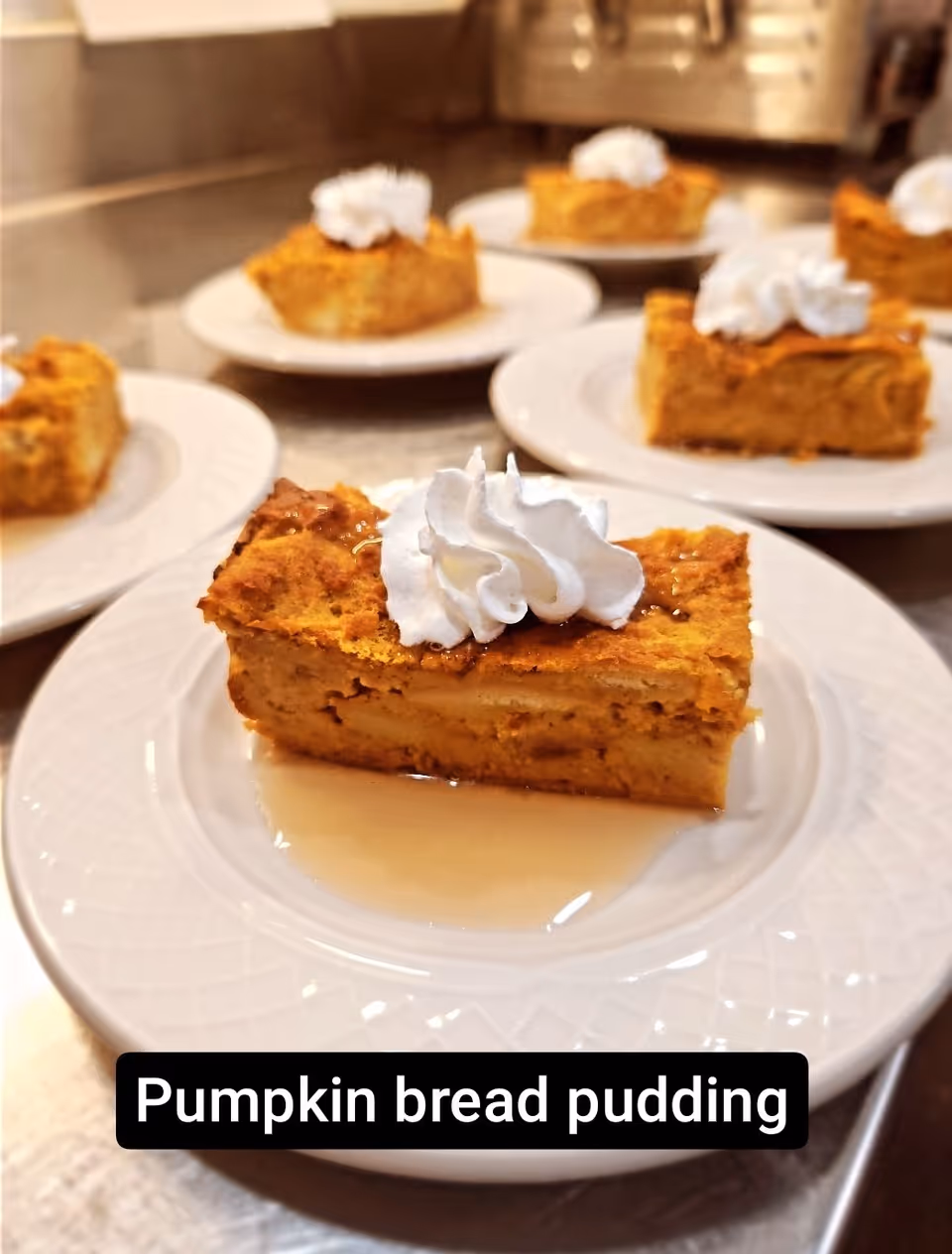 Close-up view of several plates each with a slice of pumpkin bread pudding topped with whipped cream and a drizzle of syrup, arranged on a countertop.
