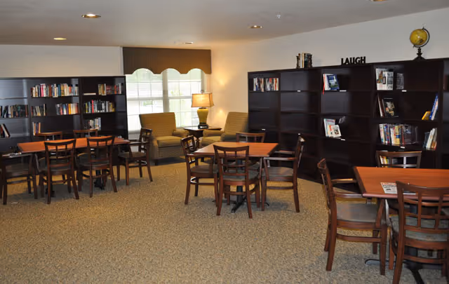 Community seating and library area with multiple tables and chairs, bookshelves, and two armchairs by a window.