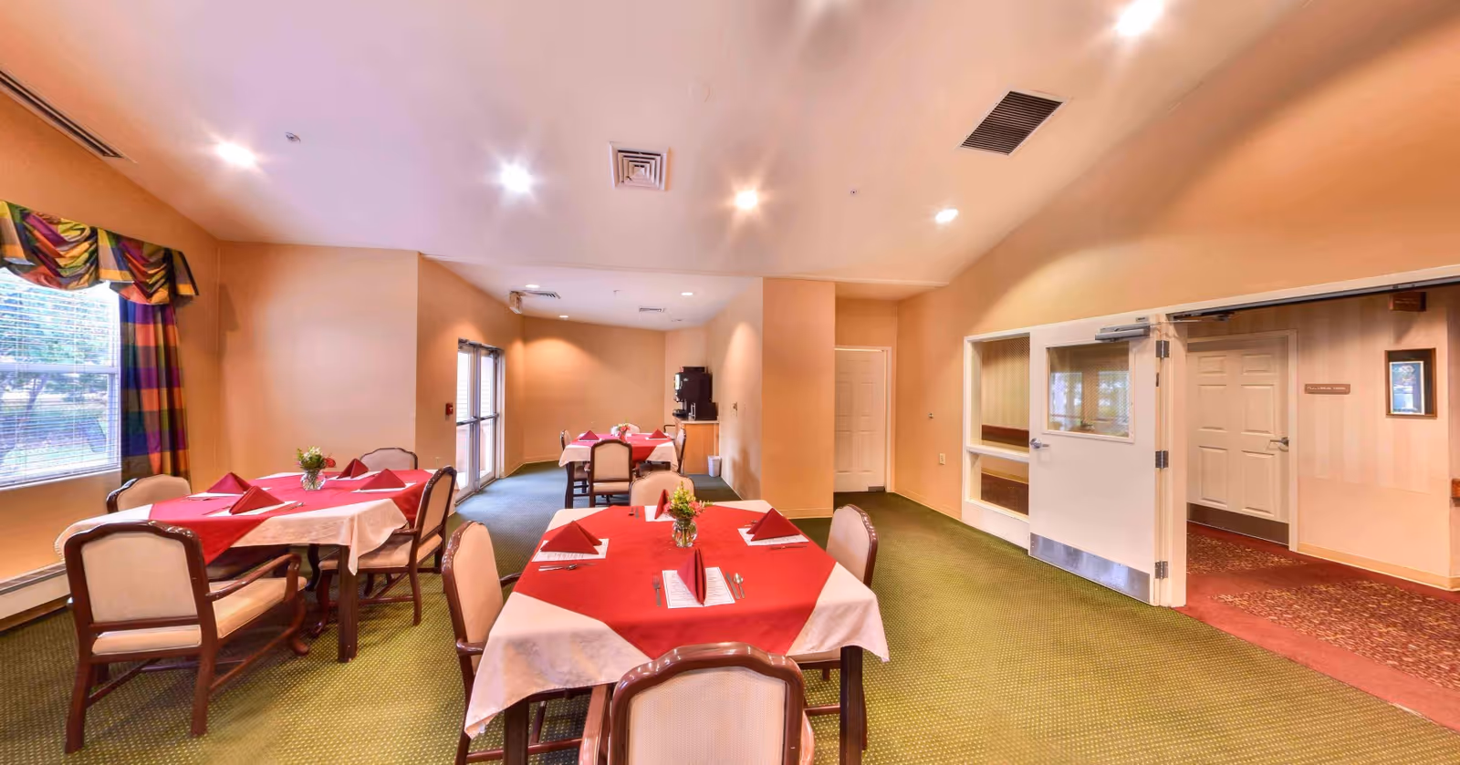 A dining room with several tables covered in red and white tablecloths, chairs, place settings, and soft overhead lighting.