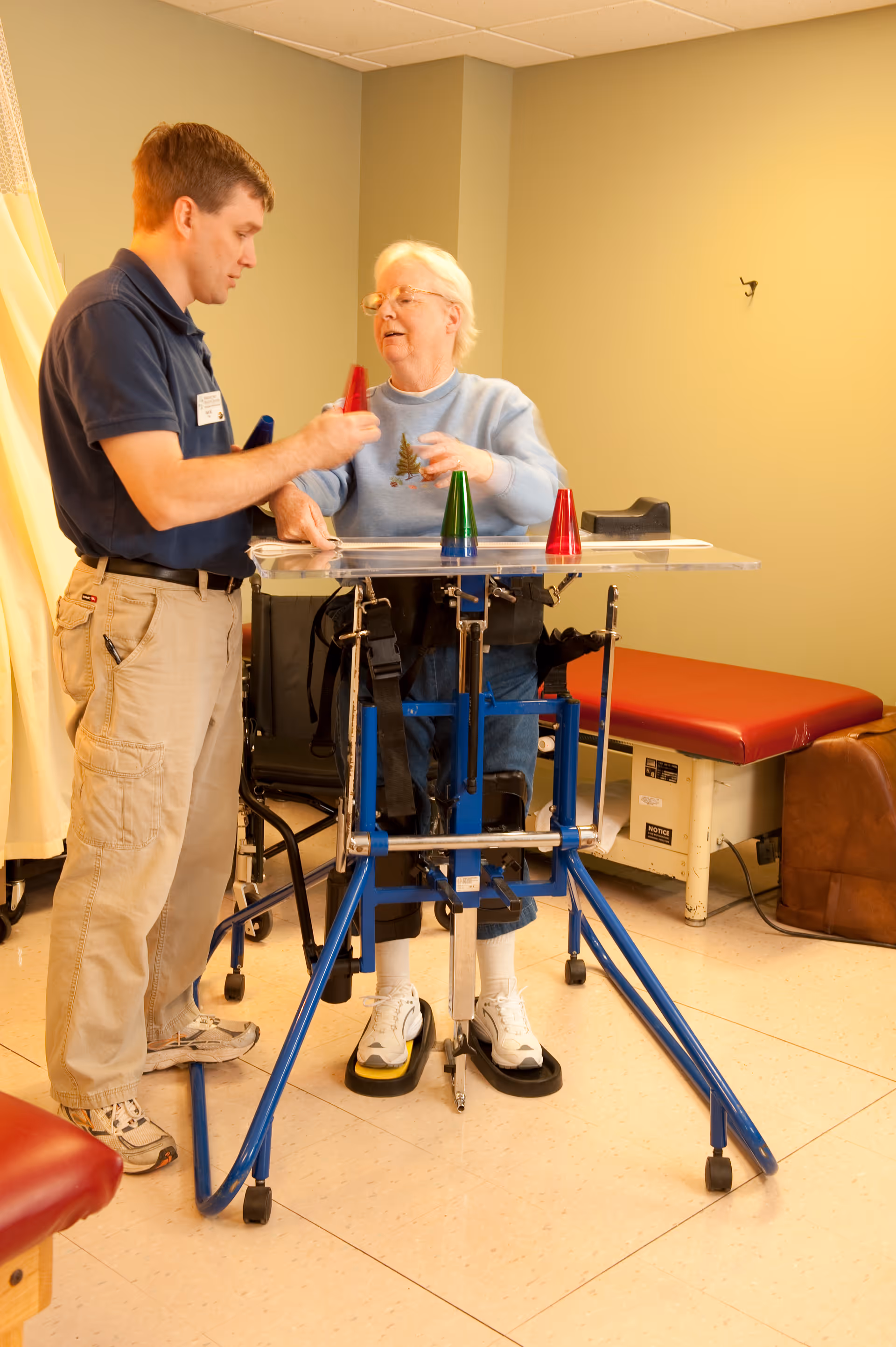 A senior woman using a standing frame device for support while engaging in a physical therapy activity with a male therapist in a clinical room. The woman is placing colored cones on a clear tray attached to the device.