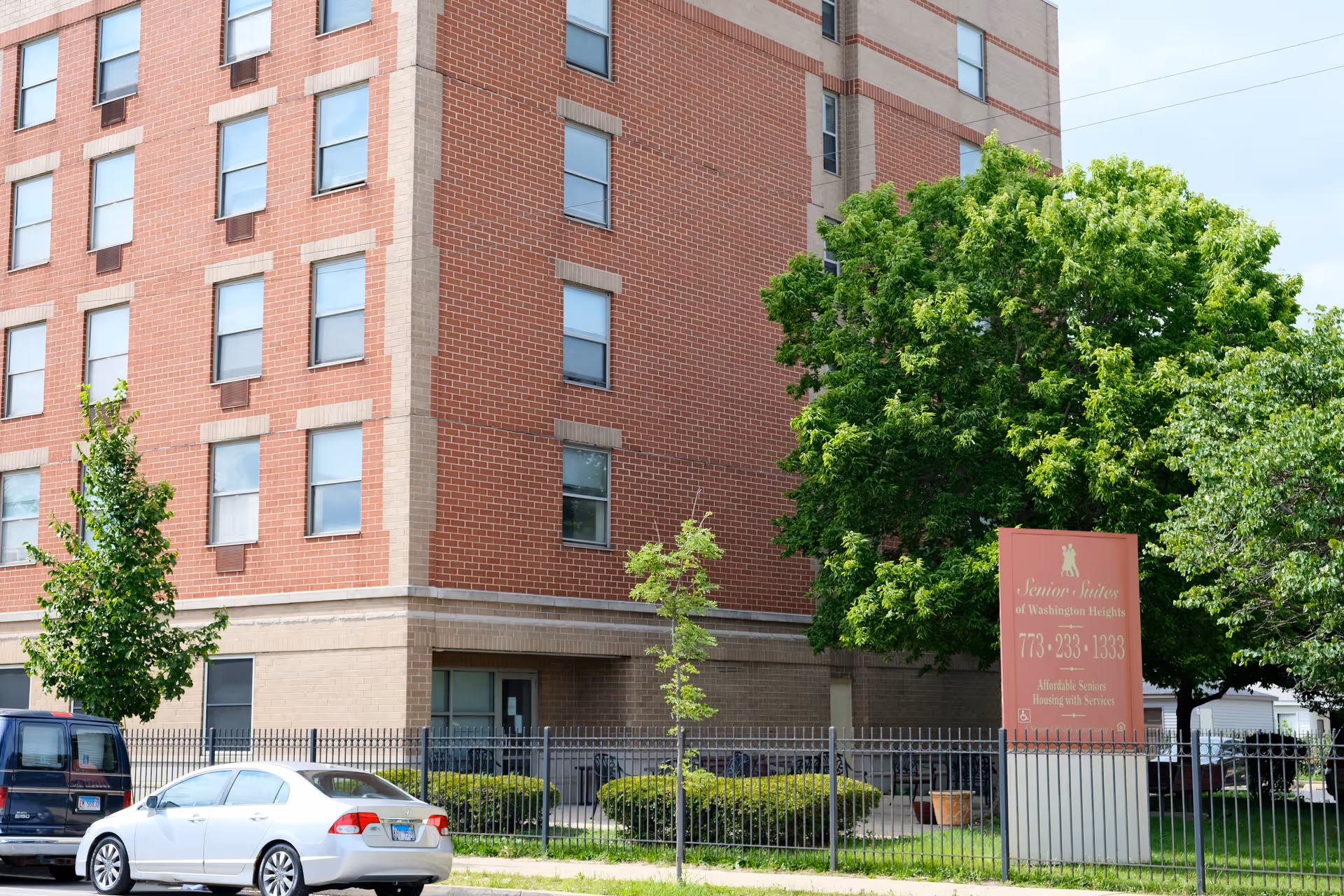 Red-brick multi-story senior living building with trees, a sign for Senior Suites of Washington Heights, and cars parked in front.