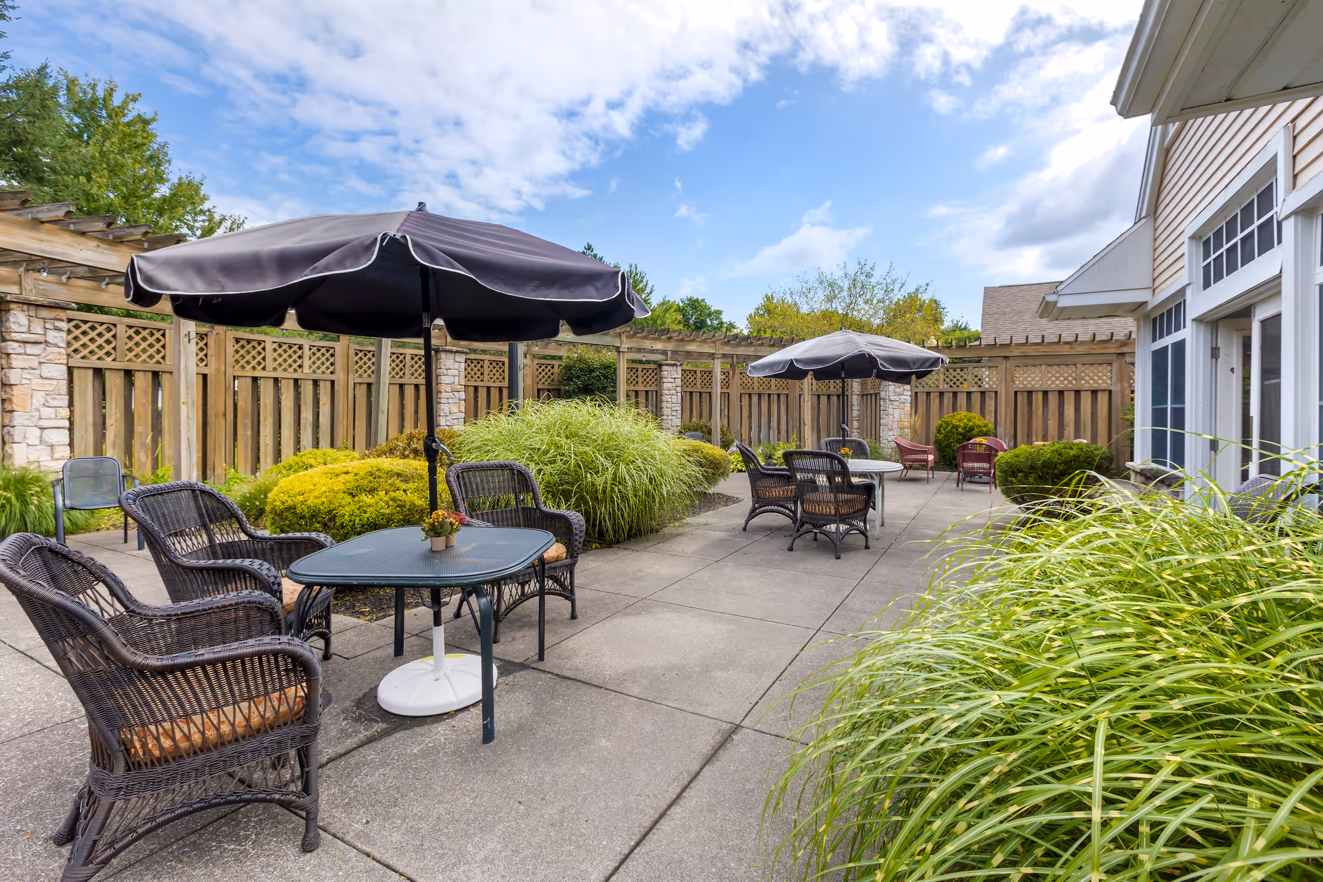 Outdoor patio area with several black wicker chairs and tables, each shaded by large black umbrellas. The patio is surrounded by green bushes and a wooden fence with stone pillars. The sky is partly cloudy with blue patches visible.