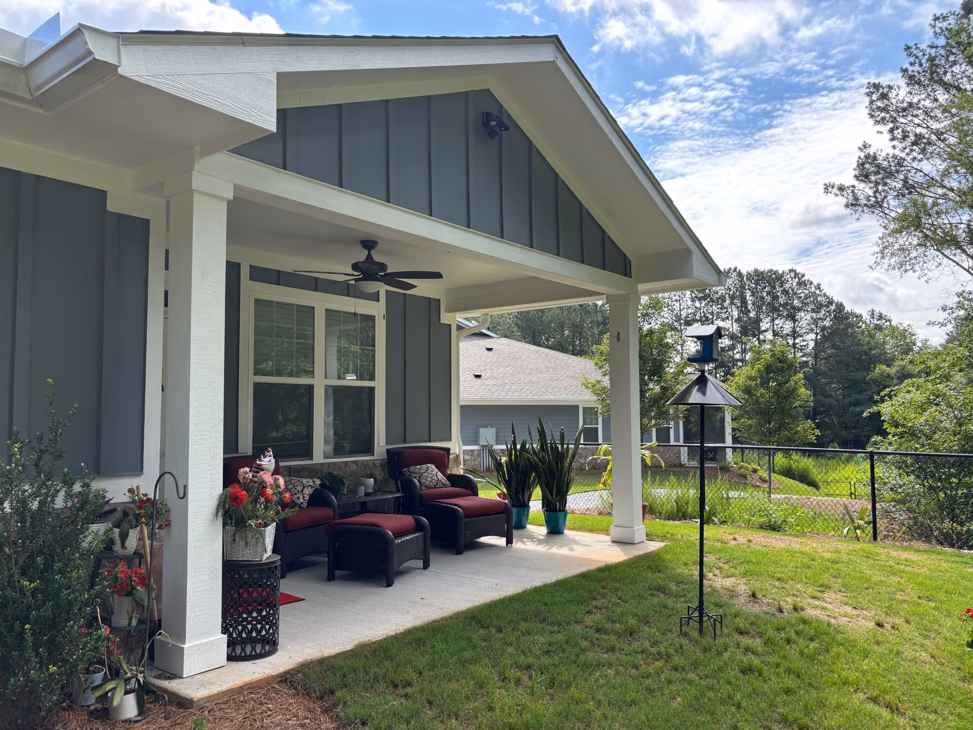 Covered outdoor patio area with two cushioned chairs and matching ottomans, a ceiling fan, potted plants, and a bird feeder on a stand in a grassy backyard with trees and a neighboring house visible in the background.