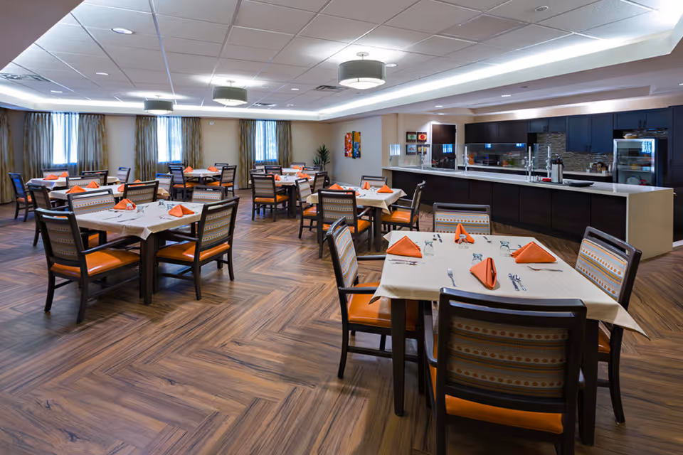 A spacious dining room with multiple tables set with white tablecloths, orange folded napkins, and silverware. The room has wooden flooring, several windows with curtains, modern ceiling lights, and a long counter area with dark cabinetry and a glass partition.