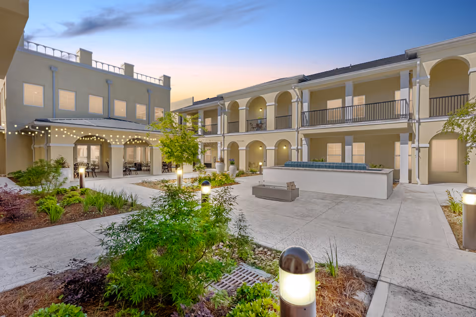 Outdoor courtyard area of a senior living facility at dusk with well-lit pathways, greenery, and a two-story building featuring balconies and arches. String lights hang over a covered seating area with tables and chairs.