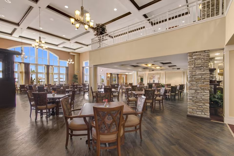 Spacious dining room in a senior living facility with multiple wooden tables and chairs arranged neatly. Large windows allow natural light to fill the room, and chandeliers hang from the coffered ceiling. Stone pillars and a balcony area are visible, creating an elegant and welcoming atmosphere.