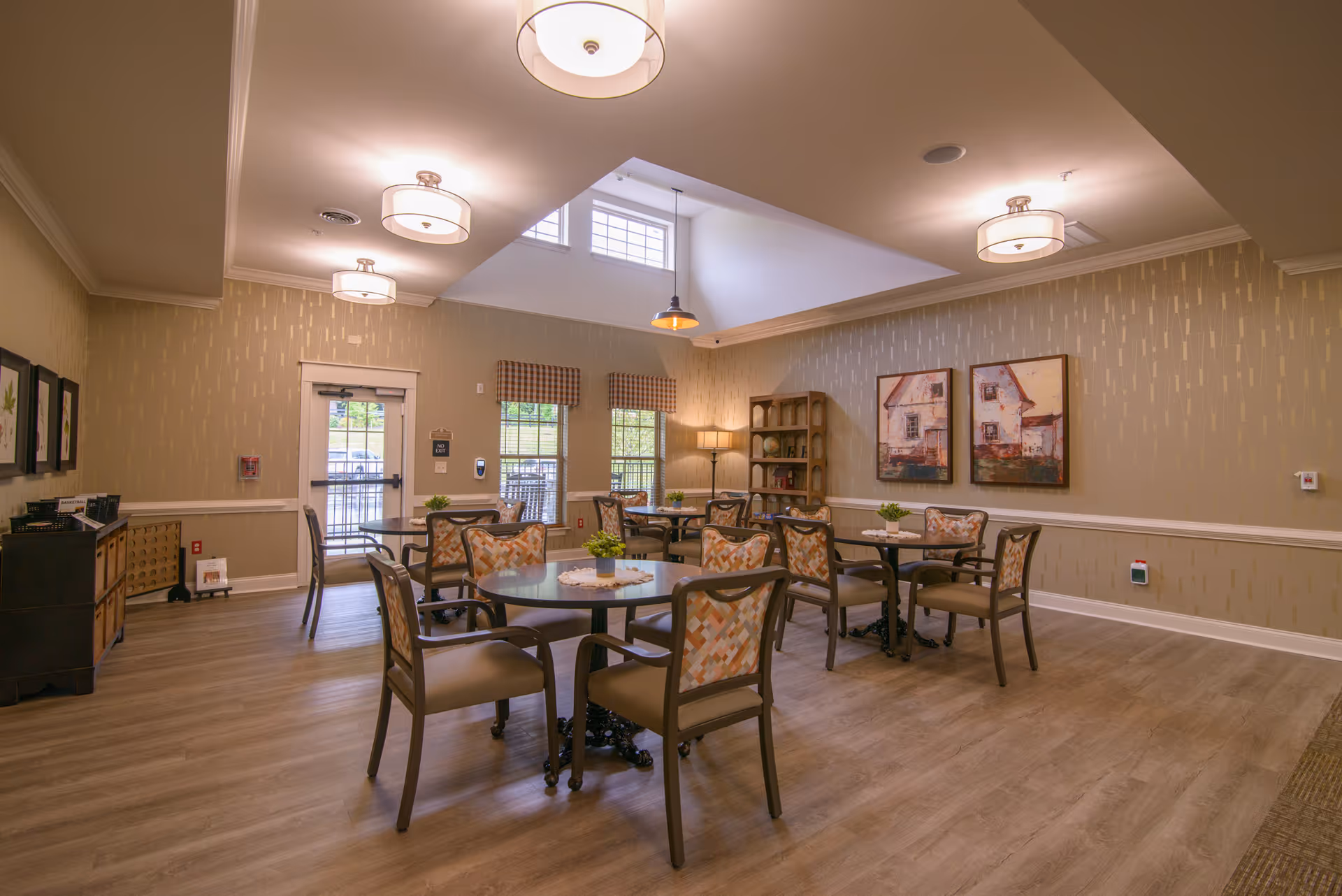 A well-lit dining room with multiple round tables and chairs arranged neatly. The room features light wood flooring, beige walls with subtle patterns, and several ceiling lights. There are two large windows with checkered valances, a door leading outside, framed artwork on the walls, and a wooden shelving unit with decorative items in the corner.