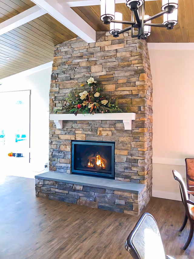 A cozy stone fireplace with a fire burning inside, topped with a white mantel decorated with a floral arrangement. The room has wooden flooring and a wooden ceiling with white beams. Part of a dining table and chairs are visible on the right side.