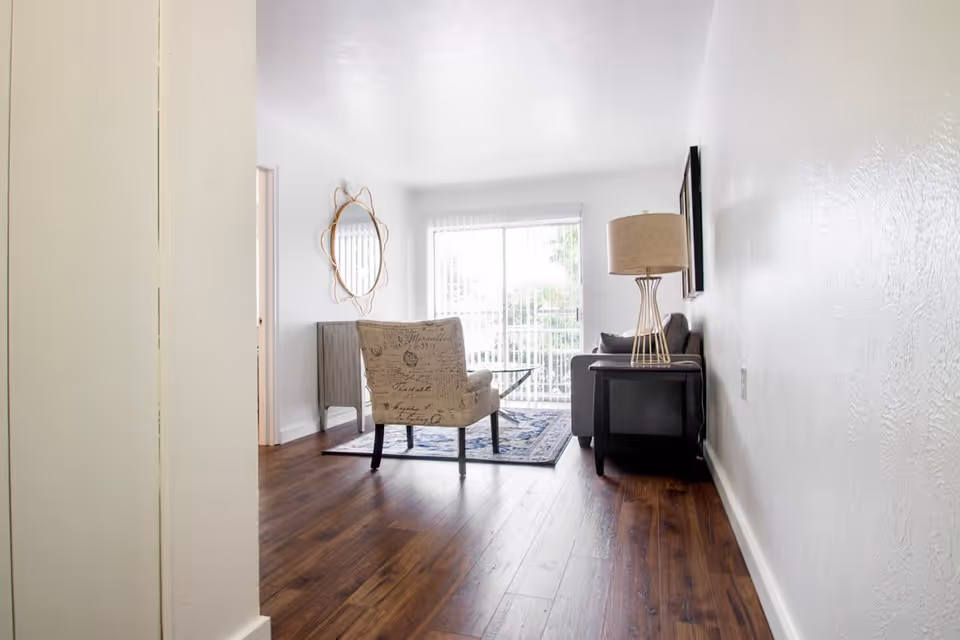 A bright living room with wooden flooring, a patterned armchair, a gray sofa, a side table with a lamp, a decorative wall mirror, and a large window with vertical blinds letting in natural light.