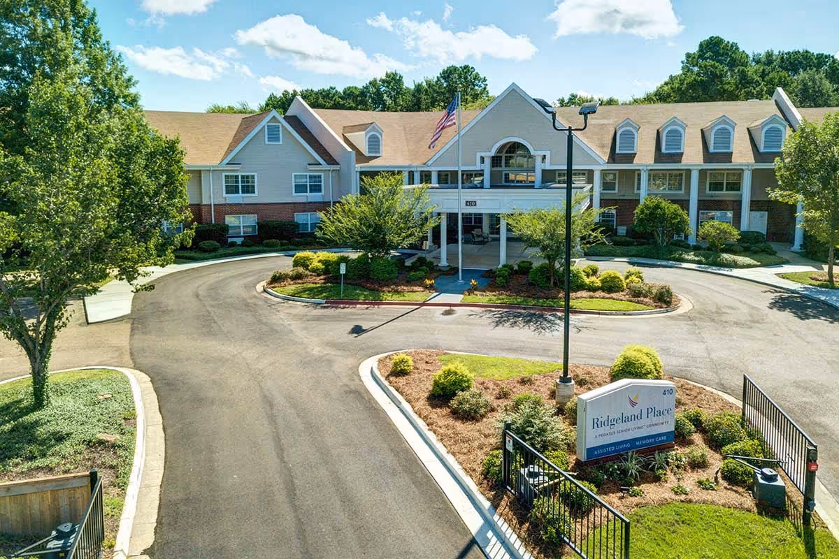 Front exterior view of Ridgeland Place, a senior living community building with a circular driveway, landscaped greenery, and an American flag flying on a flagpole in front of the entrance.