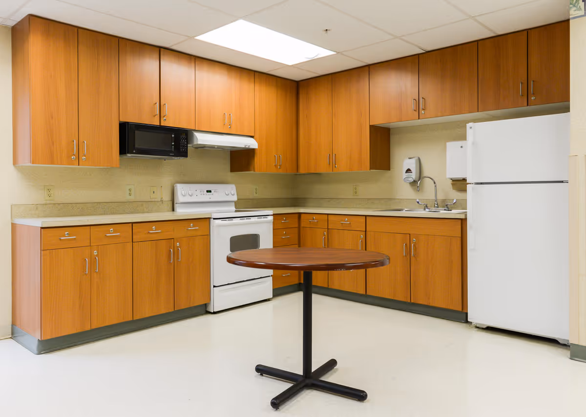 A clean kitchen area with wooden cabinets, a white stove with oven, a microwave, a white refrigerator, a sink with a soap dispenser and paper towel holder, and a round wooden table with a black metal base in the center.
