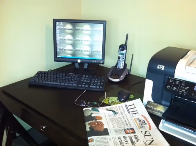 A desk setup with a computer monitor, keyboard, cordless phone, printer, eyeglasses, and a newspaper titled 'The Sun'. The desk is dark wood and placed against a light green wall.