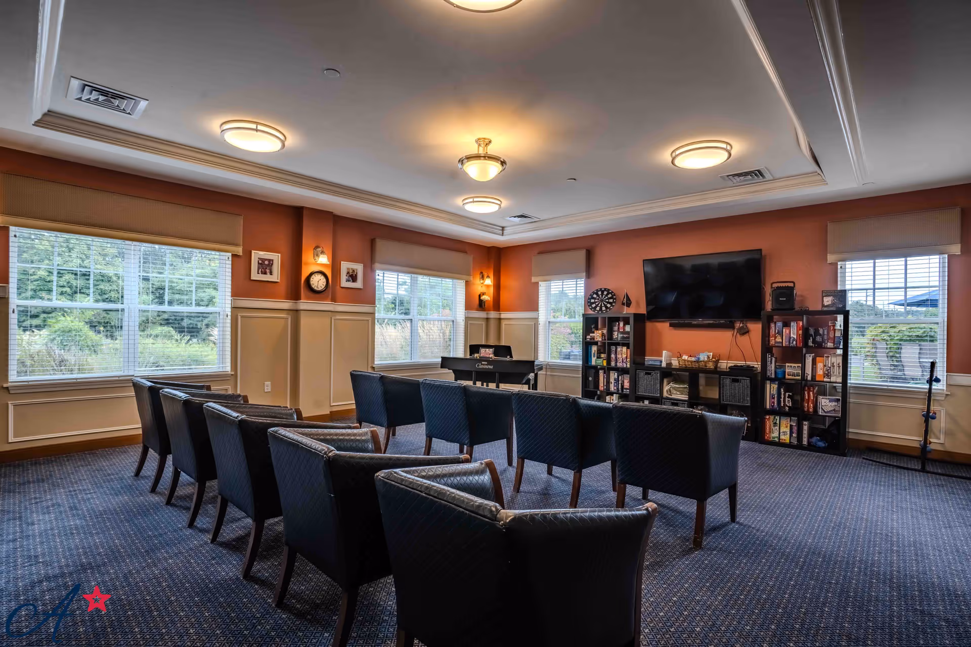 A spacious common room with multiple black cushioned chairs arranged in rows facing a wall-mounted flat screen TV. The room has large windows with beige blinds letting in natural light, a dartboard, shelves filled with board games and books, and a piano in the corner. The walls are painted in warm tones with white wainscoting, and the ceiling has recessed lighting fixtures.