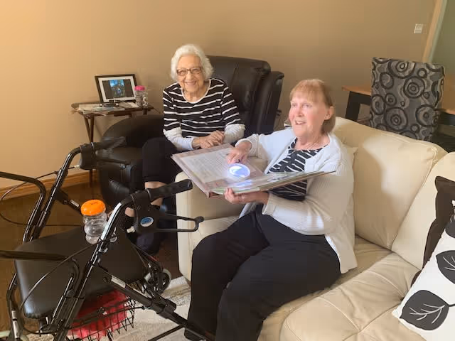 Two elderly women sitting in a living room; one woman is seated on a black recliner chair and smiling, while the other woman is seated on a cream-colored sofa holding an open photo album. A walker with an orange cup is positioned in front of them. The room has beige walls, a small side table with a framed photo, and a patterned chair in the background.