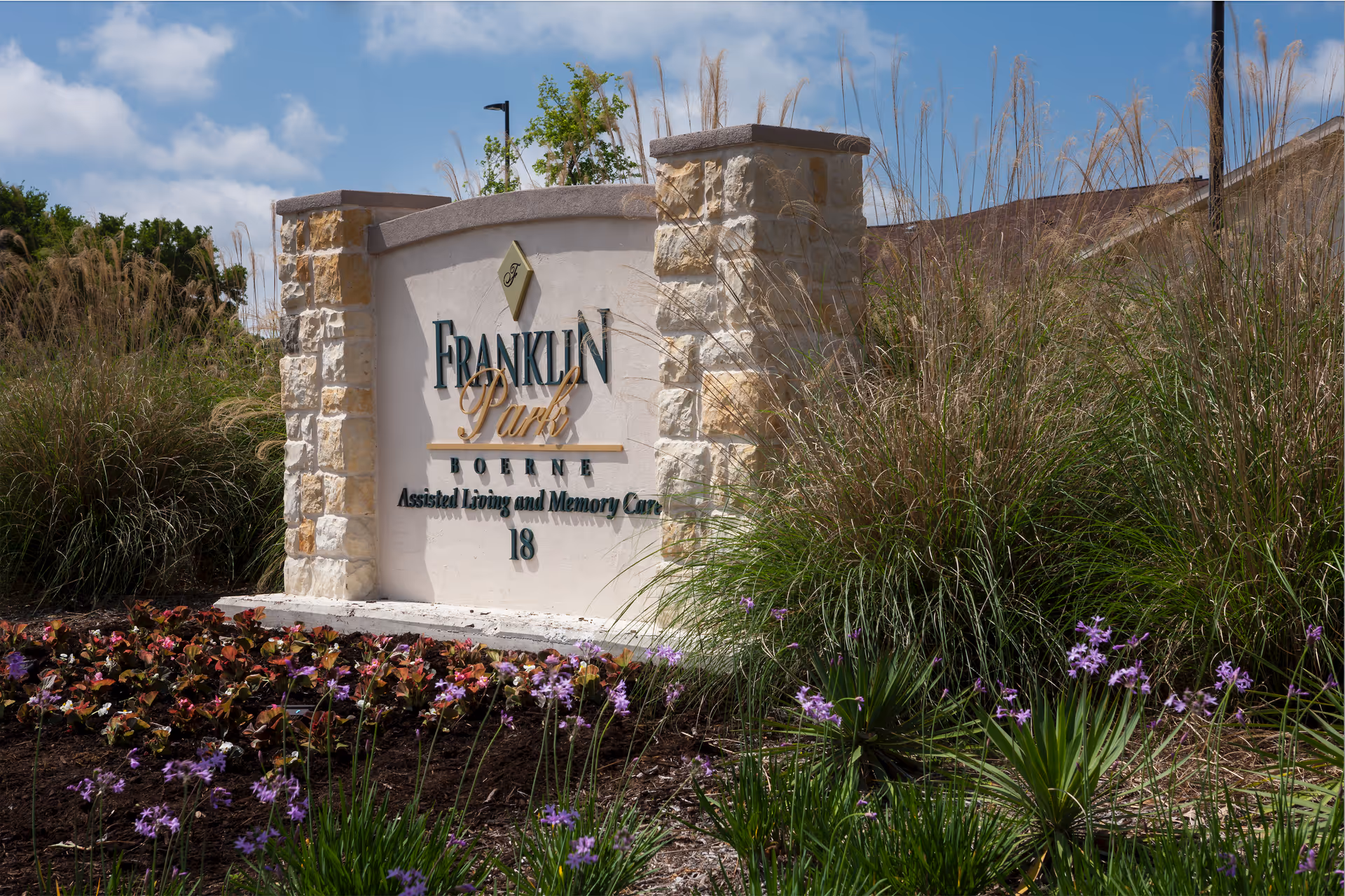Stone sign for Franklin Park Boerne Assisted Living and Memory Care surrounded by ornamental grasses and purple flowers under a partly cloudy sky.