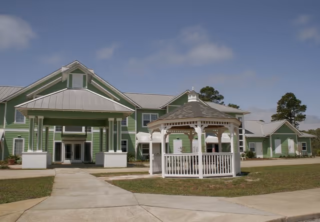 Exterior view of a green senior living facility building with a metal roof and a white gazebo in front. The building has multiple windows and a covered entrance. The sky is clear with a few clouds.