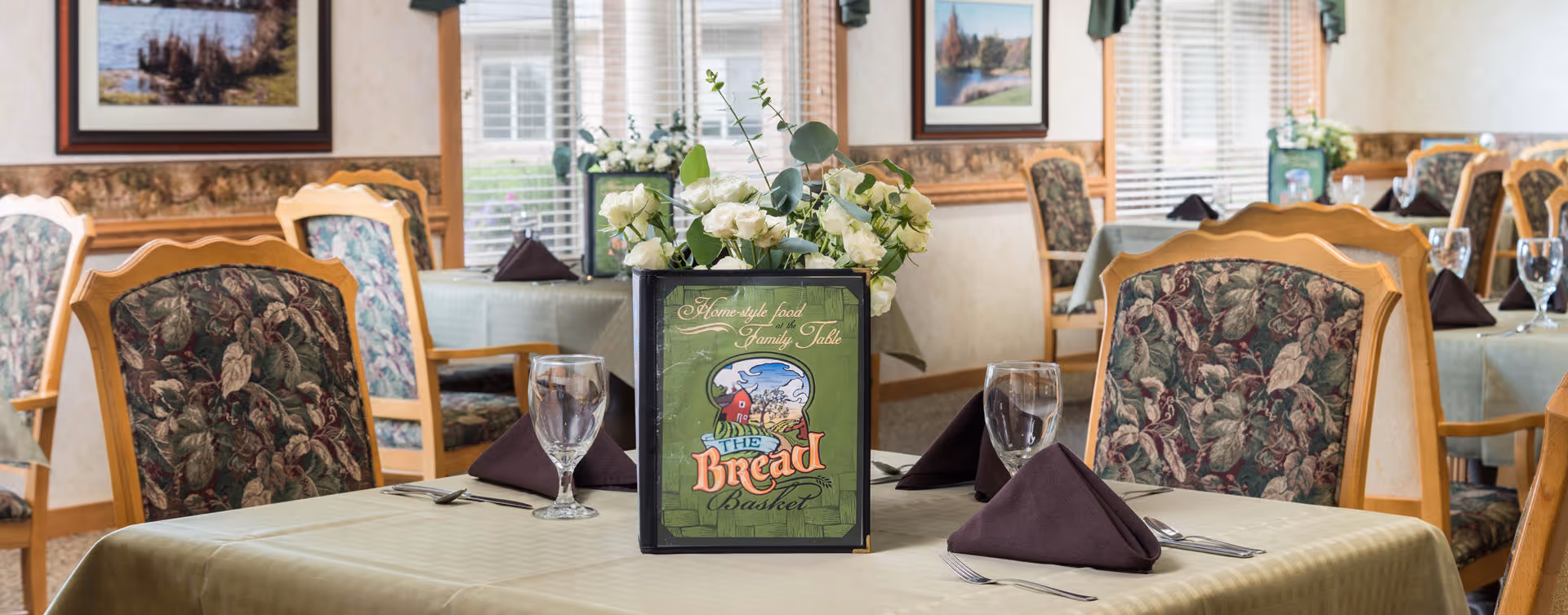 A dining room with tables covered in beige tablecloths, each set with folded dark napkins, silverware, and water glasses. The chairs have floral patterned upholstery. A centerpiece with white flowers and a menu titled 'The Bread Basket' is on the table in the foreground. The room has framed landscape paintings on the walls and windows with blinds letting in natural light.
