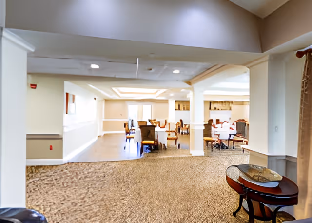 Spacious communal dining room with tables and chairs visible through an open archway in a senior living facility.