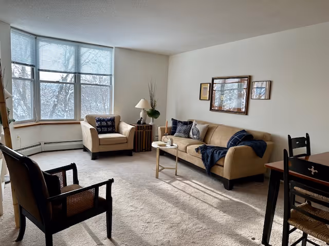 A cozy living room with a large window showing a snowy outdoor scene. The room features a beige sofa with dark blue pillows and a throw blanket, a matching armchair, a wooden side table with a lamp and decorative items, two dark wooden chairs, and a small dining table partially visible on the right. The walls are light-colored with framed artwork hanging above the sofa.
