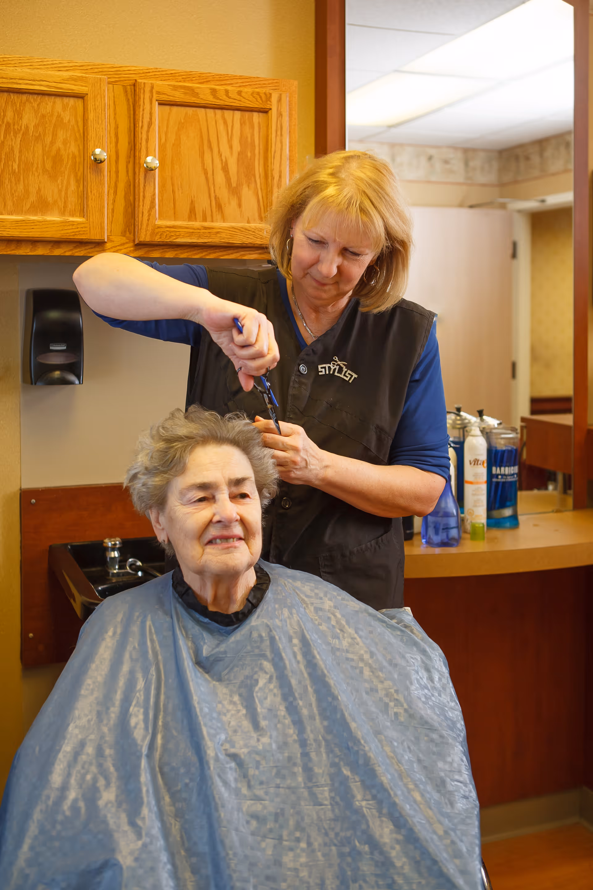 An elderly woman is getting her hair cut by a stylist in a salon area. The stylist is standing behind her, carefully trimming her hair with scissors. The elderly woman is wearing a protective cape, and there are wooden cabinets and hair care products visible in the background.