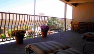 A covered outdoor patio area with cushioned lounge chairs and potted plants along a railing, overlooking a scenic view with hills in the background.