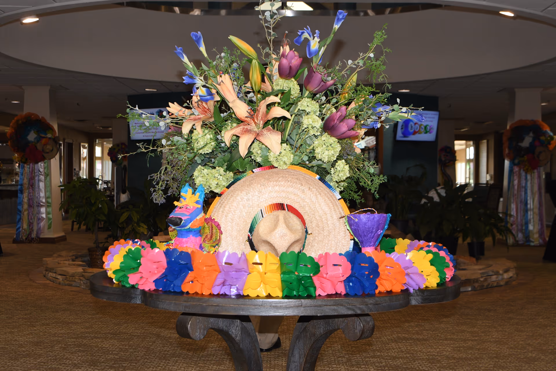 Lobby table display with a large floral arrangement, a straw sombrero and colorful paper garlands in an interior communal space.