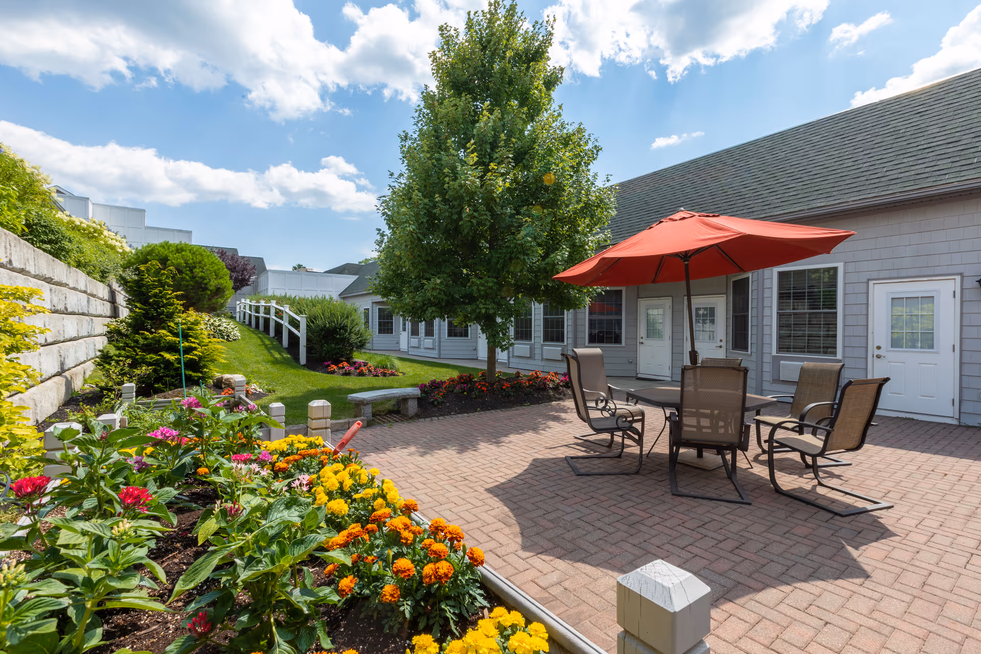Outdoor courtyard patio at a senior living facility with a table, chairs and red umbrella, flowerbeds and the building facade.