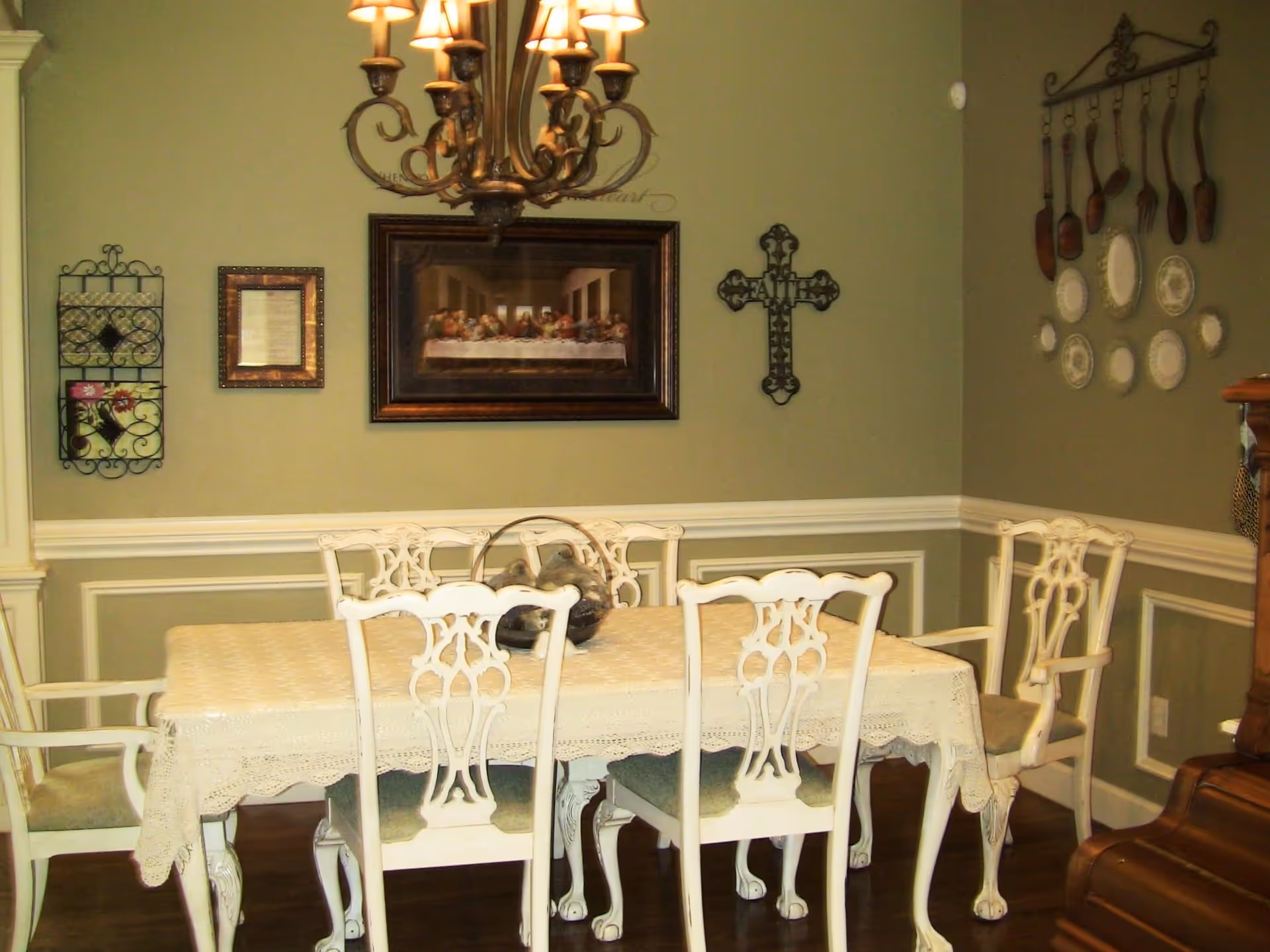 Dining room with a rectangular table covered by a lace tablecloth, six white carved chairs, a chandelier, and decorative wall art.