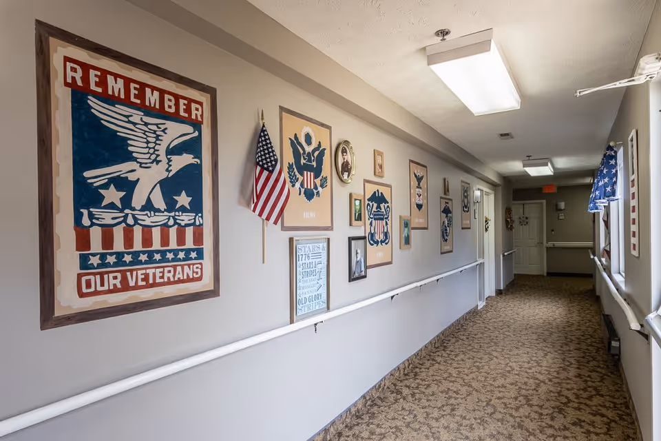 A hallway in a senior living facility decorated with patriotic-themed artwork and American flags. The walls feature framed pictures and posters honoring veterans, including one large poster that says 'Remember Our Veterans' with an eagle design. The hallway has beige walls, a patterned carpet, handrails on both sides, and ceiling lights.