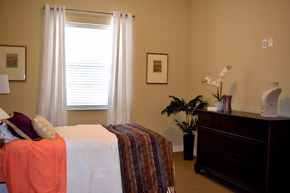 A cozy bedroom with a single bed covered in white and patterned blankets, and several pillows. There is a window with white curtains behind the bed, two framed pictures on the walls, a dark wooden dresser with decorative items including a plant and a vase, and a potted plant on the floor beside the dresser.