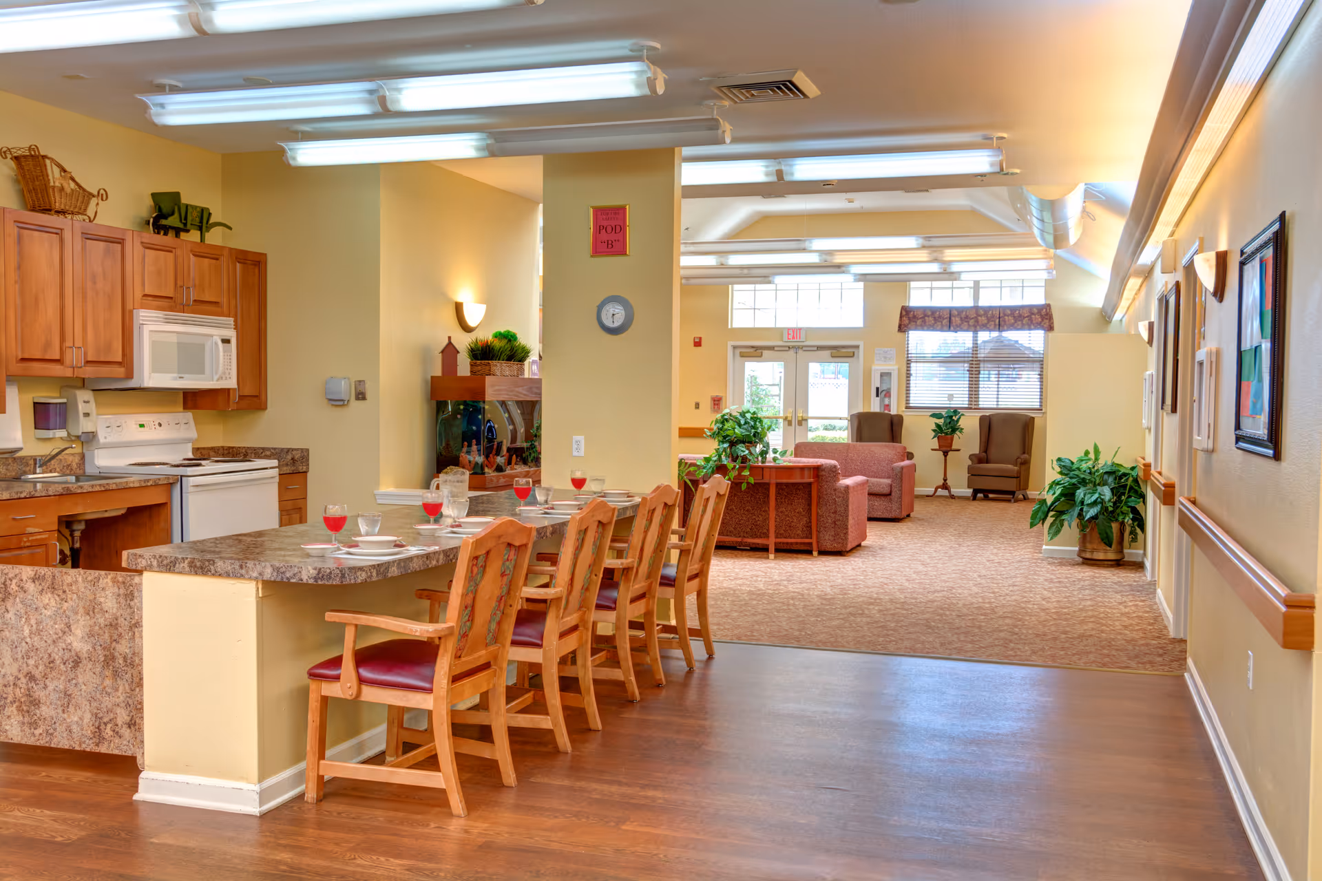 Interior view of a senior living facility showing a kitchen area with wooden cabinets, a stove, microwave, and a countertop with six chairs set with plates and glasses. Beyond the kitchen is a living room area with armchairs, sofas, plants, and large windows letting in natural light.