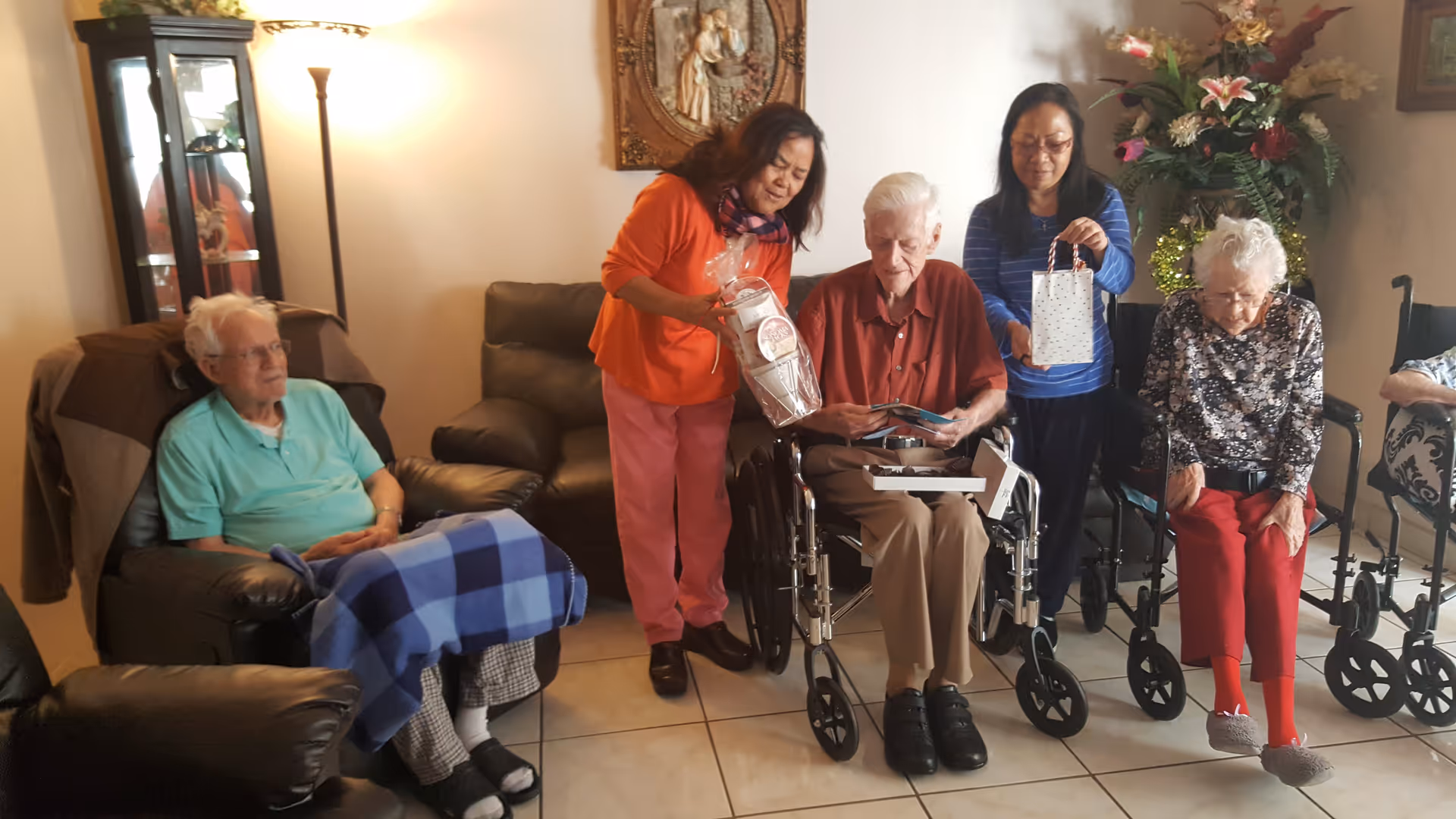 A group of elderly people and two caregivers in a cozy living room setting. One elderly man is seated in a wheelchair opening a gift box, while a caregiver in an orange top and pink pants leans over him holding a wrapped gift. Another caregiver in a blue striped top stands behind an elderly woman in a wheelchair holding a gift bag. Another elderly man is seated on a recliner with a blue checkered blanket over his legs. The room has a floor lamp, a display cabinet, and floral decorations.